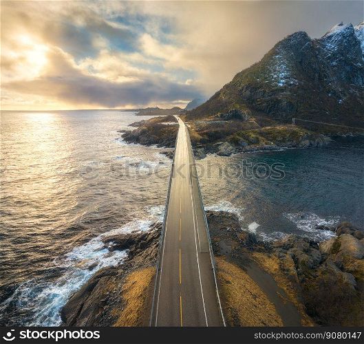 Aerial view of bridge, sea with waves and mountains at sunset in Lofoten Islands, Norway. Landscape with beautiful road, water, rocks, blue sky with clouds and golden sunlight. Top view from drone