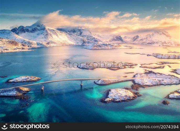Aerial view of bridge, small islands, blue sea and snowy mountains. Lofoten Islands, Norway. Fredvang bridges at sunset in winter. Landscape with rocks in snow, road, sky, clouds. Top view from drone