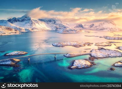 Aerial view of bridge, small islands, blue sea and snowy mountains. Lofoten Islands, Norway. Fredvang bridges at sunset in winter. Landscape with rocks in snow, road, sky, clouds. Top view from drone