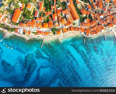 Aerial view of buildings with orange roofs, beach, clear blue sea in summer day. Vacation in beautiful Baska city, Krk island, Croatia. Top drone view of houses, streets, promenade, adriatic sea bay