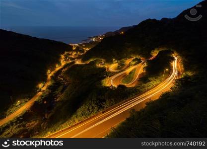 Aerial view of cars driving on curved, zigzag road or street on mountain hill with natural forest trees in rural area of New Taipei City, Taiwan at night