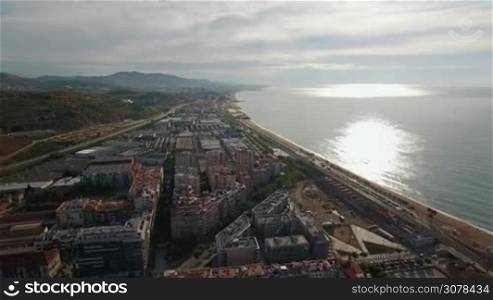 Aerial view of coast line, blue sea, railways and roofs of buildings against horizon with hills and blue cloudy sky, Barcelona, Spain