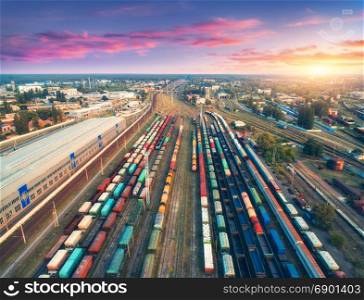 Aerial view of colorful freight trains. Railway station. Cargo trains. Wagons with goods on railroad. Heavy industry. Industrial scene with trains, city buildings and cloudy sky at sunset. Top view