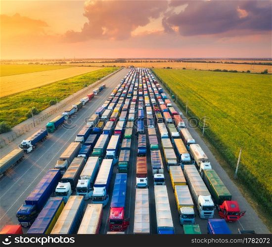 Aerial view of colorful trucks in terminal at sunset in summer. Top view of logistic center. Transportation. Cargo transport, shipping. International trucking. View from drone of trucks, road, sky