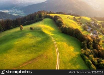 Aerial view of country road in green hills at sunset in summer. Top view from drone of rural dirt road, meadows , mountains, forest in spring. Beautiful landscape with trail, trees, grass. Slovenia