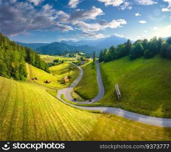 Aerial view of country road in green meadows at sunset in summer. Top drone view of rural road, alpine mountains. Colorful landscape with curved highway, hills, fields, green grass, blue sky. Slovenia