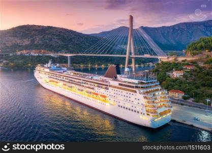 Aerial view of cruise ship and beautiful bridge at night. Port in Dubrovnik, Croatia. Summer landscape with boat, harbor, city lights, mountains at sunset. Luxury cruise. Top view of floating liner