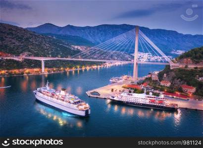 Aerial view of cruise ship at harbor and beautiful bridge at night. Landscape with ships and boats in harbour, city illumination, road, mountains, blue sea at sunset. Top view. Floating liner in port. Aerial view of cruise ship at harbor and bridge at night