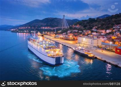 Aerial view of cruise ship at harbor at night. Landscape with ships and boats in harbour, city illumination, buildings, mountains, blue sea at sunset. Top view. Luxury cruise. Floating liner in port. Aerial view of cruise ship at harbor at night