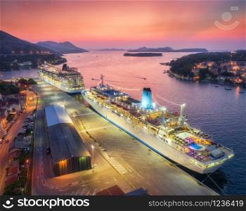 Aerial view of cruise ship in port at night in Dubrovnik. Ships and boats with illumination in harbour, city lights, buildings, mountains, blue sea, colorful sky at sunset. Top view. Luxury cruise