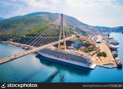 Aerial view of cruise ship under beautiful bridge at sunset. Port in Dubrovnik, Croatia. Summer landscape with road, boat, harbor, city, mountains, blue sea. Luxury cruise. Top view of floating liner