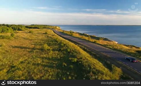 Aerial view of driving black car on the road along the cliff near the sea in summer at sunset