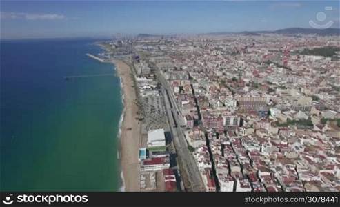 Aerial view of empty sand beach, blue sea, railways and roofs of hotels and houses against horizon and blue sky, Barcelona, Spain
