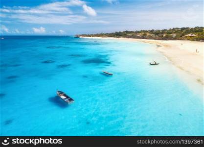 Aerial view of fishing boats and yachts on tropical sea coast with sandy beach at sunset in summer. Indian Ocean in Zanzibar. Top view of boat, palm trees, sky, clear blue water. View from above. Aerial view of boats and yachts on tropical sea coast in summer
