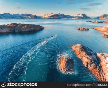 Aerial view of fishing boats, rocks in the blue sea, snowy mountains and colorful sky with clouds at sunset in winter in Lofoten islands, Norway, Landscape with two ship, blue water, waves. Top view