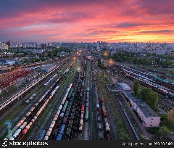 Aerial view of freight trains at colorful sunset. Railway cargo wagons on railroad. Drone view of wagons, city, pink sky with clouds at night. Depot of freight trains. Railway station. Transportation