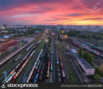 Aerial view of freight trains at colorful sunset. Railway cargo wagons on railroad. Drone view of wagons, city, pink sky with clouds at night. Depot of freight trains. Railway station. Transportation