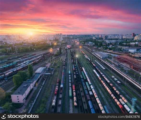 Aerial view of freight trains at colorful sunset. Railway cargo wagons on railroad. Top view of wagons, city, pink sky with clouds at night. Depot of freight trains. Railway station. Transportation