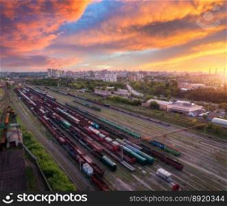 Aerial view of freight trains at sunset. Colorful railway cargo wagons on railroad. Drone view of wagons, city, sky with orange clouds. Depot of freight trains. Railway station. Industrial landscape