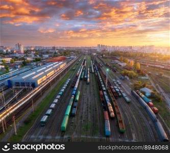 Aerial view of freight trains at sunset. Colorful railway cargo wagons on railroad. Drone view of wagons, city, sky with orange clouds. Depot of freight trains. Railway station. Industrial landscape. Aerial view of freight trains at sunset. Railway cargo wagons