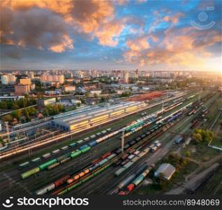 Aerial view of freight trains at sunset. Colorful railway cargo wagons on railroad. Drone view of colorful wagons, city, blue sky with clouds. Depot of freight trains. Railway station. Transportation. Aerial view of freight trains at sunset. Railway cargo wagons