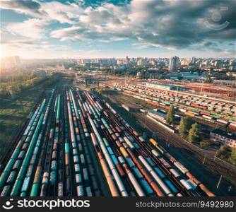 Aerial view of freight trains at sunset. Colorful railway cargo wagons on railroad. Top view of colorful wagons, city, blue sky with clouds. Depot of freight trains. Railway station. Transportation
