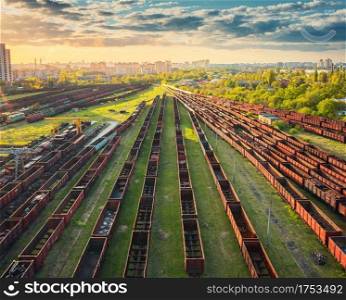 Aerial view of freight trains at sunset. Railway station. Rusty wagons on railroad. Heavy industry. Industrial landscape with train in depot, green trees, city, sky, clouds. Top view. Transportation 