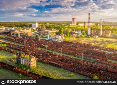 Aerial view of freight trains. Railway station with wagons. Heavy industry. Industrial landscape with train in depot, smoke stack, green trees, buildings, blue sky at sunset. Top view. Transportation