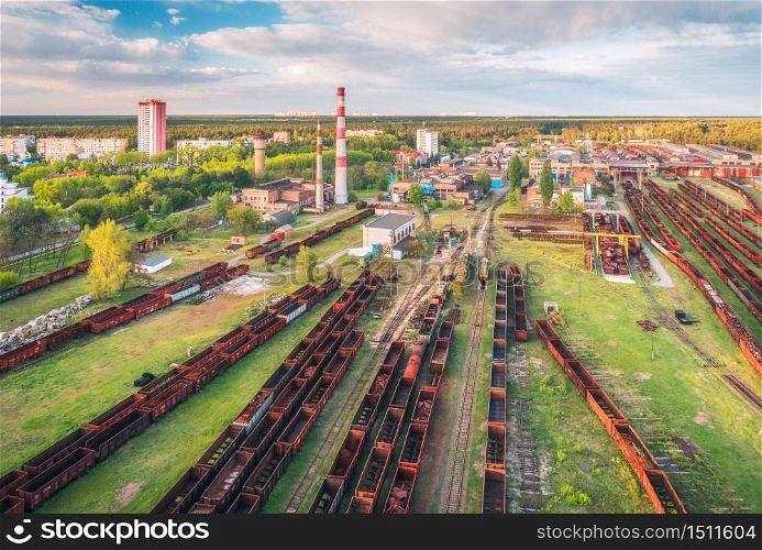 Aerial view of freight trains. Railway station with wagons. Heavy industry. Industrial landscape with train in depot, smoke stack, green trees, buildings, blue sky at sunset. Top view. Transportation