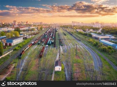 Aerial view of freight trains. Top view of railway station, wagons, railroad. Heavy industry. Industrial landscape with train in depot, green trees, buildings, colorful sky at sunset. Transportation