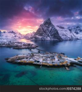 Aerial view of Hamnoy at dramatic sunset in winter in Lofoten islands, Norway. Moody landscape with blue sea, snowy mountains, rocks, village, buildings, rorbuer, road, bridge, cloudy sky. Top view