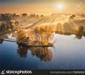 Aerial view of house with colorful trees on small island on the lake. Foggy sunrise in autumn. Beautiful landscape with village in fog, golden sunbeams, reflection in water. Fall in Ukraine. Top view