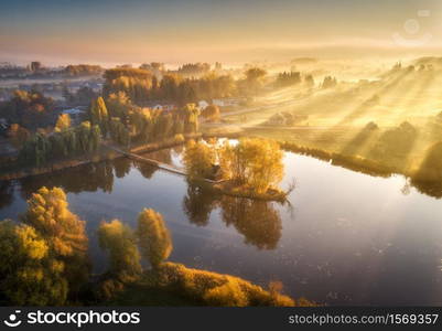 Aerial view of house with colorful trees on small island on the lake. Foggy sunrise in autumn. Beautiful landscape with village in fog, golden sunbeams, reflection in water. Fall in Ukraine. Top view