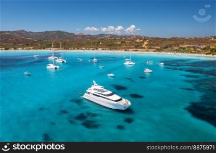 Aerial view of luxury yachts on blue sea and sandy beach at sunny day in summer. Sardinia, Italy. Aerial view of boats, yachts, sea bay, mountains, transparent water, sky. Top view. Tropical seascape