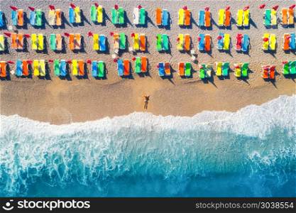 Aerial view of lying woman on the sandy beach with colorful chaise-lounges. Young woman on the sea at sunset in Europe. Top view. Landscape with girl on the seashore, blue water and waves. Resort. Aerial view of lying woman on the beach with colorful chaise-lounges