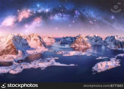 Aerial view of Milky Way arch, sea, village and snow covered mountains in winter at night. Lofoten Islands, Norway. Arctic landscape with pink starry sky, road, houses, milky way, snowy rocks. Space. Aerial view of Milky Way arch, sea, village and snowy mountains