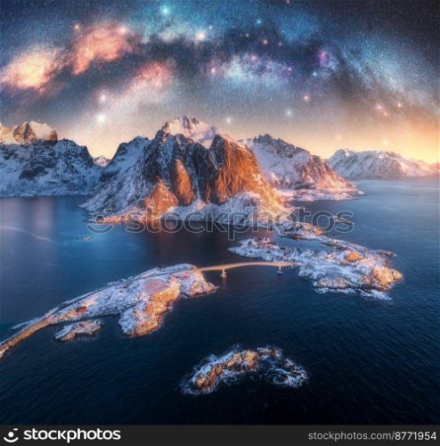 Aerial view of Milky Way arch, sea, village and snowy mountains in winter at night. Lofoten Islands, Norway. Arctic landscape with starry sky, bridge, rorbu, houses, milky way, rocks in water. Space. Aerial view of Milky Way arch, sea, village and snowy mountains