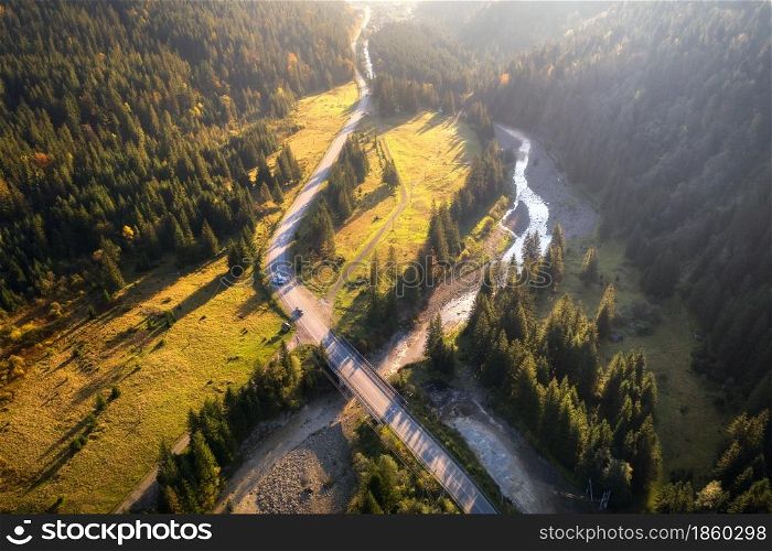 Aerial view of mountain road and beautiful forest at sunset in autumn. Top view from drone of road, bridge, woods, cars, green meadows and river. Colorful landscape with roadway in fall. Travel