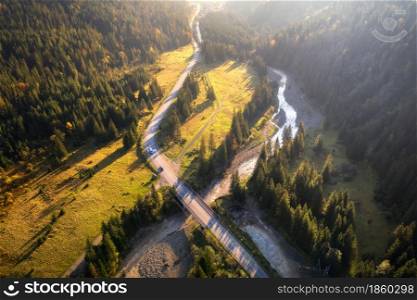 Aerial view of mountain road and beautiful forest at sunset in autumn. Top view from drone of road, bridge, woods, cars, green meadows and river. Colorful landscape with roadway in fall. Travel