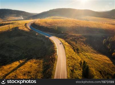 Aerial view of mountain road at sunset in autumn. Top view from drone of road in hills. Beautiful landscape with roadway, trees, green meadows, sky with golden sunlight in fall. Travel in Ukraine