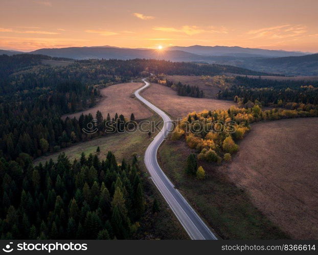 Aerial view of mountain road in autumn forest at sunset in Ukraine. Top view from drone of road in woods. Beautiful landscape with highway in hills, trees, meadows, orange sky, golden sunlight in fall. Aerial view of mountain road in autumn forest at sunset