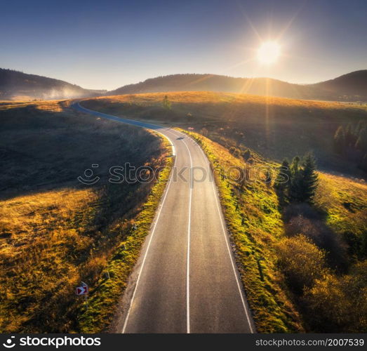 Aerial view of mountain road in forest at sunset in autumn. Top view from drone of road in woods. Beautiful landscape with roadway in hills, pine trees, green meadows, golden sunlight in fall. Travel