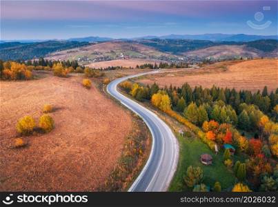 Aerial view of mountain road in forest at sunset in autumn. Top view from drone of road in woods at dusk. Beautiful landscape with roadway in hills, trees, meadows, fields, violet sky in fall. Travel