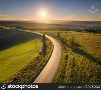 Aerial view of mountain road in green hills at sunset in autumn. Top view from drone of road. Beautiful landscape with roadway, pine trees, green fields, blue sky with orange sun in fall. Travel