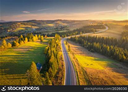 Aerial view of mountain road in village at sunset in autumn. Top view from drone of road in woods. Beautiful landscape with roadway, forest, houses, cows, green meadows, colorful sky in fall. Travel