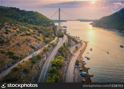 Aerial view of mountain roads and beautiful bridge at sunset. Dubrovnik, Croatia. Top view of road, boats, yachts, green trees. Summer landscape with harbor, sea coast, highway and cloudy sky