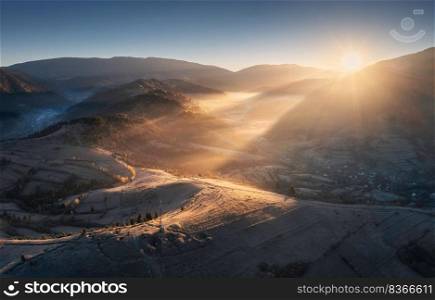 Aerial view of mountains and hills in low clouds at sunrise. Autumn in Ukraine. Top view of Carpathians mountains in fog in fall at dawn. Beautiful landscape with meadows, orange forest, village, sun. Aerial view of mountains and hills in low clouds at sunrise
