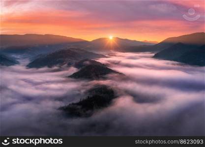 Aerial view of mountains in low clouds at sunrise in autumn. Top view of mountain hills in fog in fall at dawn. Beautiful landscape with ridges, forest, sun, colorful orange sky with pink clouds