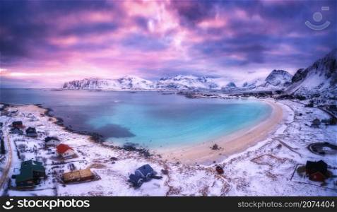 Aerial view of nordic sandy beach, sea in winter at sunset. Lofoten islands, Norway. Landscape with snowy mountains, violet sky with clouds, azure water, houses at dusk. Nature. Top view. Cold evening