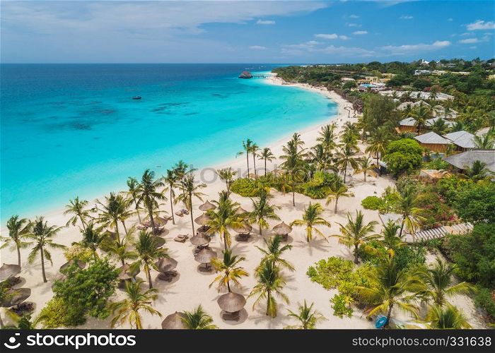 Aerial view of palms on the sandy beach of Indian Ocean at sunny day. Summer holiday in Zanzibar, Africa. Tropical landscape with palm trees, white sand, blue water, hotels. Top view of sea coast